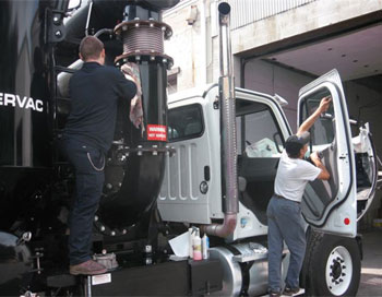 Two truck body repair technicians working servicing a white truck with its door open, parked in front of a garage.